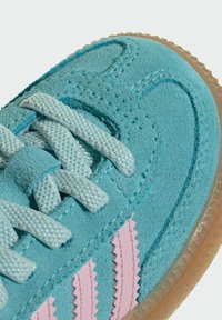 Zbliżenie na turkusowy zamszowy sneaker z jasnoniebieskimi sznurowadłami, z różowymi zygzakowatymi paskami i przezroczystą gumową podeszwą.