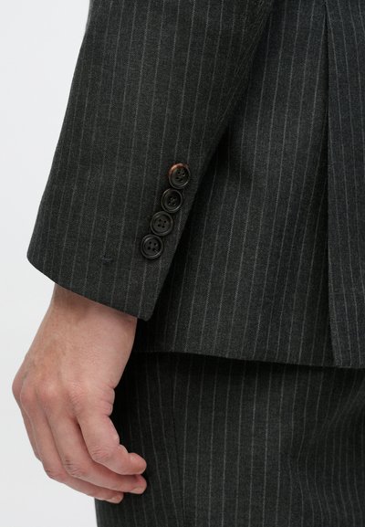Rukav sive odijelne jakne s finim prugama, s četiri crna dugmeta i mekanom teksturom tkanine. Ruka se odmara na manžeti odijela uz šav.