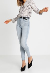 Kvinna klädd i ljusblå högmidjade skinny jeans, blus med ormskinnsmönster, svart bälte och svarta spetsiga klackar, poserar med utsträckta armar.