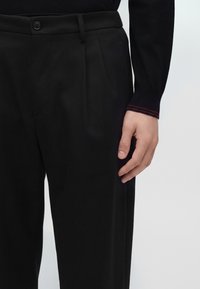 La main droite repose à côté d’un pantalon formel noir avec bouton et plis, portant un haut noir à manches longues avec des coutures rouges au poignet.