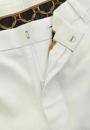 White trousers featuring a textured surface, a zip fly, and a clasp closure. Inside waistband displays a patterned fabric design.