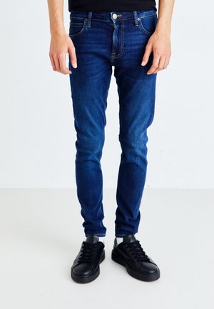 Jeans de mezclilla azul oscuro con un corte slim, que cuentan con cinco bolsillos, remaches de cobre y cierre de cremallera. Combinados con zapatillas negras.