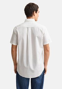 Mężczyzna w białej koszuli z krótkim rękawem na guziki i niebieskich dżinsach, zwrócony tyłem na jednolite jasne tło.