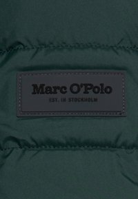 Mörkgrönt material för en vadderad jacka, med en svart rektangulär läderetikett med präglat varumärkesnamn och etableringsdetaljer. Slät textur.