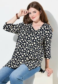 Marinebluse mit cremefarbenen Punkten, V-Ausschnitt, 3/4-Ärmel und seitlichen Schlitzen. Tragen mit hellblauen Jeans vor einem neutralen Hintergrund.