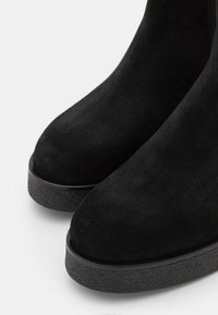 Bottines en daim noir avec bouts arrondis et semelles épaisses en caoutchouc texturé sur fond blanc.