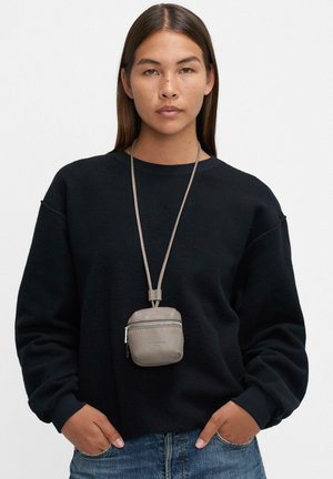 Młoda osoba o długich ciemnych włosach nosi czarną bluzę, niebieskie dżinsy oraz małą szarą saszetkę na długim naszyjniku na jednolitym tle.