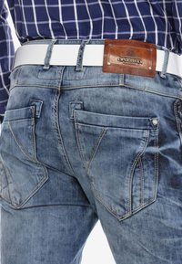 Vervaagde blauwe denim jeans beschikken over dubbele patchzakken, contrasterende stiksels en een bruine leren merklabel met metalen accenten. Witte riem zichtbaar.
