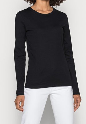 Maglietta nera a maniche lunghe in tessuto elasticizzato con scollo rotondo. Vestibilità aderente e design semplice. Indossata con pantaloni bianchi.