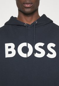 Felpa blu navy con tasca a marsupio frontale, dotata di un grande logo bianco "BOSS", lacci con puntali in argento e un tessuto testurizzato.