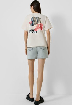 Kobieta ubrana w beżowy t-shirt z grafiką nadmorskich namiotów i homara, jasne dżinsowe szorty oraz czarne sandały na platformie, stojąca przy białej ścianie.