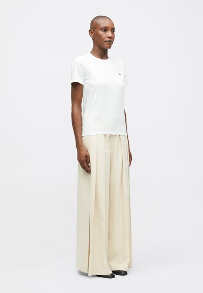 Žena s krátkými vlasy, oblečená v bílém triku s krátkým rukávem a širokých béžových skládacích kalhotách, stojící na čistém bílém pozadí.