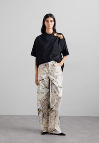 Tricou negru supradimensionat cu un design asimetric unic și un accent circular auriu, asociat cu pantaloni largi cu model în culori estompate.