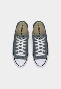 Γκρι καμβαδένια αθλητικά χαμηλού ύψους με λευκά κορδόνια, λαστιχένια μύτη και αντίθετη μαύρη επένδυση γύρω από τη σόλα. Ορατή η σήμανση "Converse All Star".