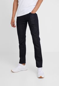 Jean en denim foncé à coupe droite, avec poches avant et coutures minimales, associé à des chaussures de sport blanches.