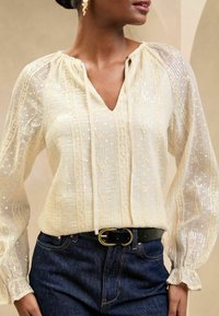 Light beige blouse with sheer fabric, intricate embroidery, and sequins. V-neckline and gathered sleeves. Paired with dark blue jeans and a black belt.