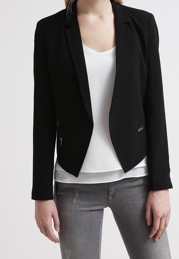 Femme portant un blazer noir cintré sur un haut blanc à col en V et un jean gris déchiré, debout devant un fond uni.