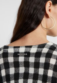 Schwarzer und weißer karierter Stricksweater mit rundem Ausschnitt und einem Etikett an der Rückseite. Große, runde goldene Creole sichtbar.