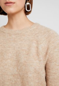 Maglione beige con collo rotondo, texture morbida e superficie leggermente pelosa. Presenta maniche raglan. Orecchini geometrici grandi visibili.