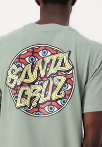 T-shirt de algodão verde com um gráfico circular com fundo vermelho, olhos azuis e texto "SANTA CRUZ" em letras estilizadas amarelas e pretas.