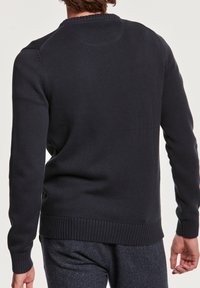Homme portant un pull en tricot bleu marine foncé et un pantalon foncé, vu de dos sur un fond uni.