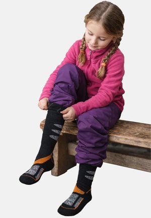 Kind met paarse broek en een roze fleecevest met rits, zwarte sokken met grijze en oranje accenten aantrekkend, zittend op een houten bankje.