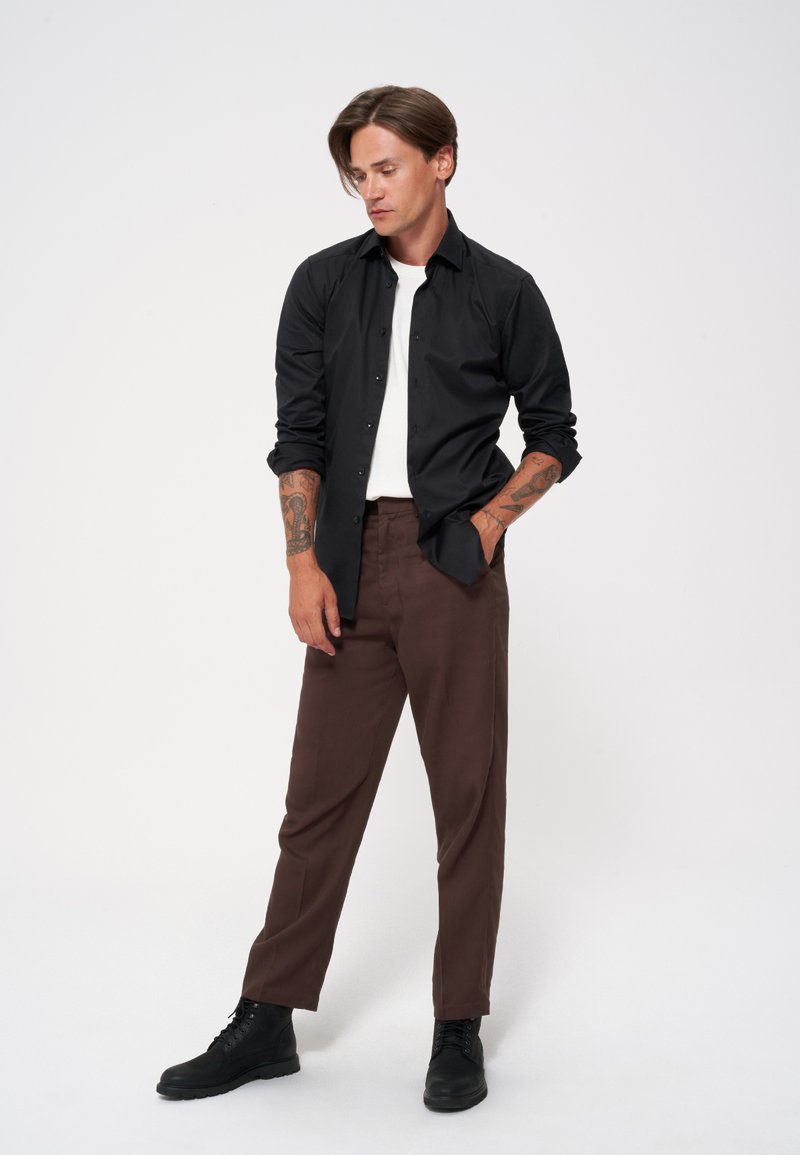 Czarna koszula z guzikami, biała koszulka z okrągłym dekoltem, brązowe spodnie dopasowane, czarne botki do kostki. Gładki materiał, dopasowany krój, widoczne tatuaże.