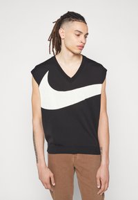 Schwarzer ärmelloser Pullover mit großem weißen Nike-Swoosh-Design, V-Ausschnitt und geripptem Saum. Modell trägt beige Hose und zeigt ein Armtattoo.