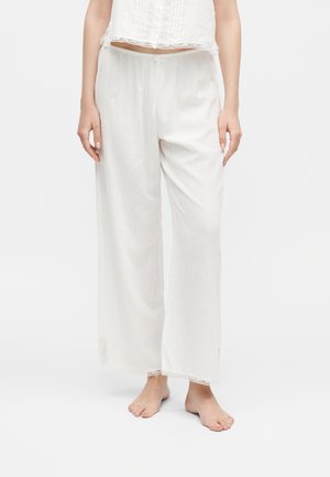 Mujer descalza con pantalones blancos de lino sueltos con cintura elástica y bordes de encaje en los puños y el dobladillo de la parte superior.