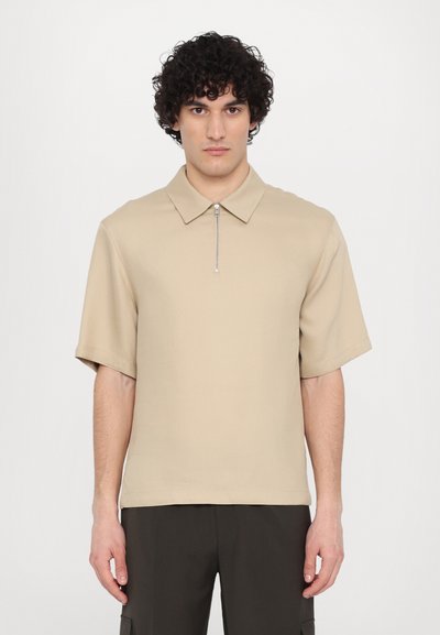 Polo de culoare bej, cu mânecă scurtă și guler cu fermoar, având o textură netedă, asociat cu pantaloni negri. Design simplu și croială confortabilă.