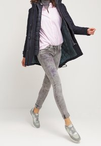 Jeans ajustados de rayas grises, camisa rosa de botones y una chaqueta acolchada azul marino. Zapatillas grises con detalles plateados. Modelo en movimiento.
