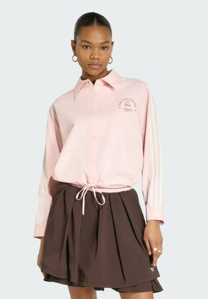 Giovane donna che indossa una camicia rosa chiaro con bottoni e logo e una gonna plissettata marrone scuro, in piedi davanti a uno sfondo semplice.