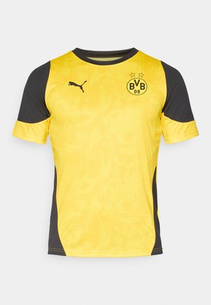 Camiseta deportiva amarilla con detalles en negro. Presenta un cuello redondo, mangas cortas y un patrón de puntos. Muestra el logotipo y el emblema de la marca.