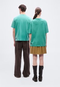 Ένας άνδρας και μια γυναίκα στέκονται πλάτη με πλάτη φορώντας ταιριαστά πράσινα μπλουζάκια· ο άνδρας φορά καφέ παντελόνι και η γυναίκα μια κίτρινη καρό φούστα με μαύρες μπότες.