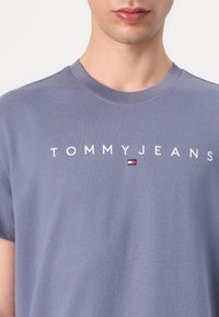 Szürke pamut póló rövid ujjakkal; a "TOMMYJEANS" felirat és egy kis színes logó díszítés jellemzi, sima textúrával.
