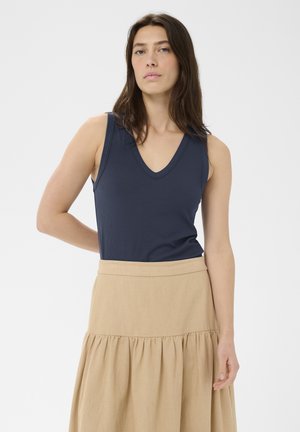 Kobieta z długimi brązowymi włosami w granatowym, bezrękawnym topie z dekoltem w serek i beżowej marszczonej spódnicy, stojąca na tle białego tła.