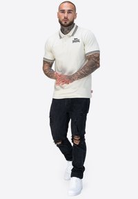 Cremfarbendes Polohemd mit schwarz-weiß gestreiftem Kragen, kombiniert mit schwarzen, zerrissenen Jeans und weißen Sneakers. Verfügt über ein Logo auf der Brust.