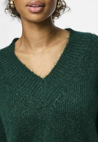 Zaļš adīts džemperis ar dziļu V-veida kakla izgriezumu, ribotu faktūru un mīkstu, pūkainu materiālu. Akcentēts ar plūstošu spīdumu.