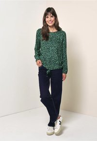 Grüne Bluse mit einem gepunkteten Muster, langen Ärmeln und einem geknoteten Vorderteil. Kombiniert mit dunkelblauen Hosen und weißen Turnschuhen mit grünen Akzenten.