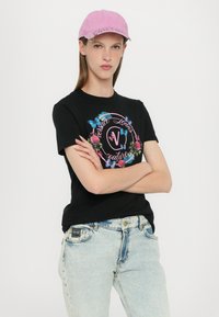 Svart t-skjorte med fargerik blomstermotiv og sommerfugltrykk, med rundt logo; brukt med lyseblå jeans og en rosa caps.