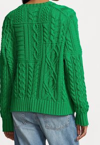 Pull vert en maille torsadée avec des motifs texturés variés, porté avec un jean denim bleu clair, vu de dos.