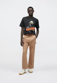 Zwart katoenen T-shirt met kleurrijk grafisch ontwerp, gecombineerd met lichtbruine katoenen broek en witte sneakers met een gumzool. Model staat ontspannen.