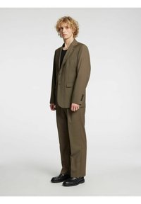 Bruin pak gemaakt van gestructureerde stof, met een colbertjas met één knoop en broek met rechte pijpen. Zwarte schoenen maken de outfit compleet.