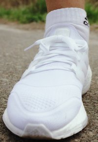 Scarpa sportiva bianca con superiore in rete texturizzata, lacci piatti e suola in gomma. Include una linguetta con logo sul tallone e un morbido polsino alla caviglia.