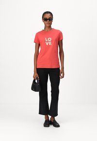 Koralle T-Shirt mit "LOVE"-Textgrafik in Creme, kombiniert mit schwarzen, weit geschnittenen Hosen mit dezentem Strukturmuster und schwarzen Slippern.