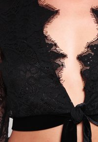 Top in pizzo nero con scollatura a V profonda, caratterizzato da bordi sfrangiati e una cintura in velluto annodata. Il pizzo presenta motivi floreali e dettagli intricati.