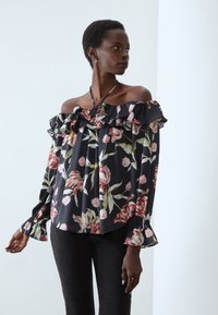Off-the-shoulder blouse in black fabric with large pink and cream floral print. Features ruffled neckline and cuffs, and slightly loose fit.