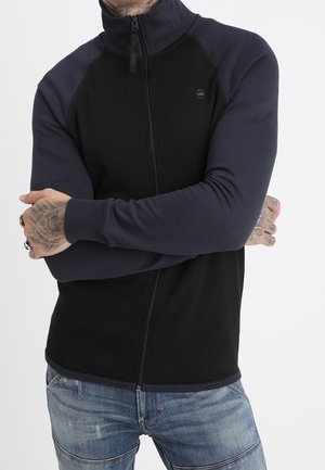 Mann mit tätowierten Händen, der eine schwarze und dunkelblaue Jacke mit Reißverschluss und blaue Jeans trägt, die Arme vor der Brust verschränkt.