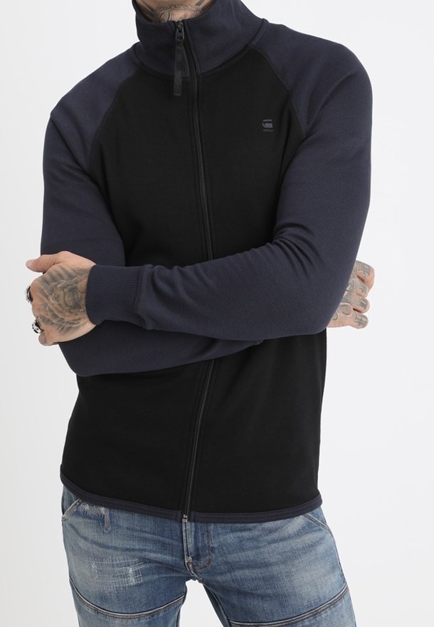 Homme aux mains tatouées portant une veste zippée noire et bleu marine foncé et un jean bleu, les bras croisés sur la poitrine.