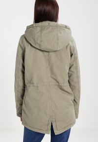 Osoba s hnědými vlasy, která má na sobě světlou olivově zelenou mikinu s kapucí a modré džíny, otočená zády k jednobarevnému pozadí.